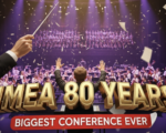 A viral, hyper-realistic YouTube thumbnail with a cinematic concert hall atmosphere. The background is the packed Grand Wayne Convention Center stage filled with hundreds of young musicians performing under dramatic purple and gold spotlights with subtle confetti falling. The composition uses a dramatic low-angle shot to focus on the main subject: a gleaming conductor's baton raised triumphantly in the foreground with sheet music pages flying around it. The image features massive 3D typography with strict hierarchy: The Primary Text reads exactly: 'IMEA 80 YEARS'. This text is massive, the largest element in the frame, rendered in polished gold chrome with glowing edges to look like a high-budget 3D render. The Secondary Text reads exactly: 'BIGGEST CONFERENCE EVER'. This text is significantly smaller, positioned below the main text with a bold red sticker-style outline and slight shadow effect. The text materials correspond to the story's concept. Crucial Instruction: There is absolutely NO other text, numbers, watermarks, or subtitles in this image other than these two specific lines. 8k, Unreal Engine 5, cinematic render
