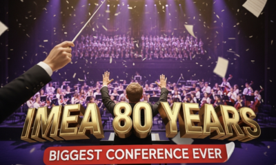 A viral, hyper-realistic YouTube thumbnail with a cinematic concert hall atmosphere. The background is the packed Grand Wayne Convention Center stage filled with hundreds of young musicians performing under dramatic purple and gold spotlights with subtle confetti falling. The composition uses a dramatic low-angle shot to focus on the main subject: a gleaming conductor's baton raised triumphantly in the foreground with sheet music pages flying around it. The image features massive 3D typography with strict hierarchy: The Primary Text reads exactly: 'IMEA 80 YEARS'. This text is massive, the largest element in the frame, rendered in polished gold chrome with glowing edges to look like a high-budget 3D render. The Secondary Text reads exactly: 'BIGGEST CONFERENCE EVER'. This text is significantly smaller, positioned below the main text with a bold red sticker-style outline and slight shadow effect. The text materials correspond to the story's concept. Crucial Instruction: There is absolutely NO other text, numbers, watermarks, or subtitles in this image other than these two specific lines. 8k, Unreal Engine 5, cinematic render