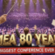 A viral, hyper-realistic YouTube thumbnail with a cinematic concert hall atmosphere. The background is the packed Grand Wayne Convention Center stage filled with hundreds of young musicians performing under dramatic purple and gold spotlights with subtle confetti falling. The composition uses a dramatic low-angle shot to focus on the main subject: a gleaming conductor's baton raised triumphantly in the foreground with sheet music pages flying around it. The image features massive 3D typography with strict hierarchy: The Primary Text reads exactly: 'IMEA 80 YEARS'. This text is massive, the largest element in the frame, rendered in polished gold chrome with glowing edges to look like a high-budget 3D render. The Secondary Text reads exactly: 'BIGGEST CONFERENCE EVER'. This text is significantly smaller, positioned below the main text with a bold red sticker-style outline and slight shadow effect. The text materials correspond to the story's concept. Crucial Instruction: There is absolutely NO other text, numbers, watermarks, or subtitles in this image other than these two specific lines. 8k, Unreal Engine 5, cinematic render