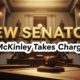 A viral, hyper-realistic YouTube thumbnail with a Legislative News atmosphere. The background is a blurred, majestic interior of the Indiana Statehouse with warm, golden lighting. The composition uses a Low Angle to focus on the main subject: a detailed Faceless Gavel resting on a wooden desk. The image features massive 3D typography with strict hierarchy: The Primary Text reads exactly: 'NEW SENATOR'. This text is massive, the largest element in the frame, rendered in Gold to look like a high-budget 3D render. The Secondary Text reads exactly: 'McKinley Takes Charge'. This text is significantly smaller, positioned below the main text. It features a thick, distinct white border/outline (sticker style) to contrast against the background. Make sure text 2 is always different theme, style, effect and border compared to text 1.