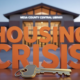 A viral, hyper-realistic YouTube thumbnail with a Business/Community atmosphere. The background is a blurred, high-quality shot of the Mesa County Central Library exterior at twilight with warm, inviting lighting. The composition uses a slightly Low Angle to focus on the main subject: a detailed 3D House Key resting on a wooden table. The image features massive 3D typography with strict hierarchy: The Primary Text reads exactly: 'HOUSING CRISIS'. This text is massive, the largest element in the frame, rendered in Matte Orange to look like a high-budget 3D render. The Secondary Text reads exactly: 'SPEAK UP TONIGHT'. This text is significantly smaller, positioned below the main text. It features a thick, distinct white border/outline (sticker style) to contrast against the background. Make sure text 2 is always different theme, style, effect and border compared to text 1.