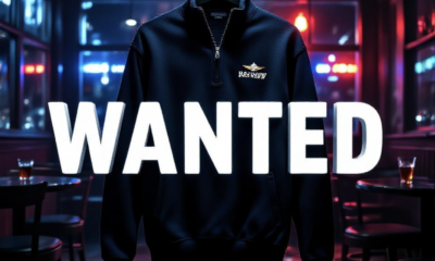 A viral, hyper-realistic YouTube thumbnail with a dark, tense crime atmosphere. The background is the dimly lit interior of a Colorado bar at night with scattered chairs, spilled drinks, and red-blue police lights flashing through windows. The composition uses a dramatic low angle to focus on the main subject: a black quarter-zip Navy pullover hanging ominously in the foreground, the U.S. Navy emblem glowing under harsh spotlight. The image features massive 3D typography with strict hierarchy: The Primary Text reads exactly: 'WANTED'. This text is massive, the largest element in the frame, rendered in cold brushed steel with red