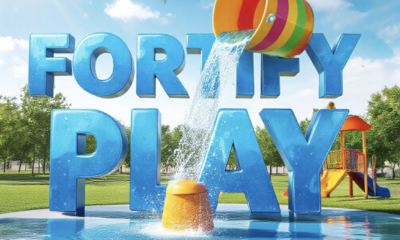 A viral, hyper-realistic YouTube thumbnail with a bright, summer community vibe atmosphere. The background is a sunny, green park environment with a modern splash pad and playground equipment under a clear blue sky with warm sunlight. The composition uses a low angle to focus on the main subject: a colorful, 3D-rendered bucket dumping water (from a splash pad feature). The image features massive 3D typography with strict hierarchy: The Primary Text reads exactly: 'FORTIFY PLAY'. This text is massive, the largest element in the frame, rendered in glossy blue water texture to look like a high-budget 3D render. The Secondary Text reads exactly: '$1.5M GOAL'. This text is significantly smaller, positioned below the main text. It features a thick, distinct white border/outline (sticker style) to contrast against the background. Make sure text 2 is always different theme, style, effect and border compared to text 1.