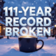 A viral, hyper-realistic YouTube thumbnail with a cozy winter storm atmosphere. The background is downtown Grand Junction buried in fresh snow at golden hour, warm coffee shop windows glowing against the cold blue dusk with snowflakes gently falling. The composition uses a low-angle shot looking up to focus on the main subject: a massive steaming coffee mug planted in deep snow like a victory trophy. The image features massive 3D typography with strict hierarchy: The Primary Text reads exactly: '111-Year Record Broken'. This text is massive, the largest element in the frame, rendered in frosted ice-blue chrome with fresh snow accumulating on the letters to look like a high-budget 3D render. The Secondary Text reads exactly: 'Grand Junction'. This text is significantly smaller, positioned below the main text with a thick orange glowing border like warm coffee shop light to contrast against the cold background. The text materials correspond to the story's concept. Crucial Instruction: There is absolutely NO other text, numbers, watermarks, or subtitles in this image other than these two specific lines. 8k, Unreal Engine 5, cinematic render