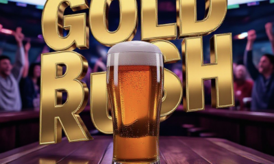 A viral, hyper-realistic YouTube thumbnail with a energetic Sports Bar atmosphere. The background is a bustling, modern pub interior with blurred cheering fans and multiple TV screens glowing with hockey game footage. The composition uses a Low Angle to focus on the main subject: a foaming glass of golden beer resting on a wooden bar counter. The image features massive 3D typography with strict hierarchy: The Primary Text reads exactly: 'GOLD RUSH'. This text is massive, the largest element in the frame, rendered in Gold Chrome to look like a high-budget 3D render. The Secondary Text reads exactly: 'RECORD OPENING'. This text is significantly smaller, positioned below or next to the main text. It features a thick, distinct color border/outline (sticker style) to contrast against the background. Make sure text 2 is always different theme, style, effect and border compared to text 1.