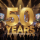 A viral, hyper-realistic YouTube thumbnail with a warm, golden Western Colorado atmosphere. The background is the packed interior of Grand Junction Convention Center filled with bright vendor booths, Colorado mountain murals, and smiling crowds under dramatic overhead lighting. The composition uses a wide, slightly low angle to focus on the main subject: a massive gleaming golden house model with "50 YEARS" carved into it. Image size should be 3:2. The image features massive 3D typography with strict hierarchy: The Primary Text reads exactly: '50 YEARS'. This text is massive, the largest element in the frame, rendered in polished gold chrome with subtle light flares to look like a high-budget 3D render. The Secondary Text reads exactly: 'GRAND JUNCTION EXPO'. This text is significantly smaller, positioned below the main text with a thick red sticker-style outline and slight drop shadow to pop against the background. The text materials correspond to the story's concept. Crucial Instruction: There is absolutely NO other text, numbers, watermarks, or subtitles in this image other than these two specific lines. 8k, Unreal Engine 5, cinematic render