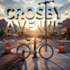 A viral, hyper-realistic YouTube thumbnail with a vibrant urban renewal atmosphere. The background is a sunlit Grand Junction street under construction with orange barrels, fresh asphalt, and Colorado red rock mountains in the distance with dramatic golden hour lighting. The composition uses a low-angle shot to focus on the main subject: a gleaming chrome bicycle locked to a brand-new LED streetlight pole emerging from smooth fresh pavement. Image size should be 3:2. The image features massive 3D typography with strict hierarchy: The Primary Text reads exactly: 'CROSBY AVENUE'. This text is massive, the largest element in the frame, rendered in polished chrome metal with realistic reflections to look like a high-budget 3D render. The Secondary Text reads exactly: 'GETS BIKE LANES'. This text is significantly smaller, positioned below the main text. It features a thick neon green outline with glowing edge effect to contrast against the background. Make sure text 2 is always different theme, style, effect and border compared to text 1.