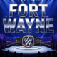 A viral, hyper-realistic YouTube thumbnail with a high-energy wrestling arena atmosphere. The background is the packed Allen County War Memorial Coliseum at night with thousands of phone lights glowing and blue-purple WWE lighting sweeping across the crowd. The composition uses a dramatic low-angle shot to focus on the main subject: a massive, gleaming WWE Championship belt floating above the ring. Image size should be 3:2. The image features massive 3D typography with strict hierarchy: The Primary Text reads exactly: 'FORT WAYNE'. This text is massive, the largest element in the frame, rendered in polished chrome with electric blue glow edges to look like a high-budget 3D render. The Secondary Text reads exactly: 'MAY 23'. This text is significantly smaller, positioned below the main text. It features a thick red outline with white fill and subtle fire sparks to contrast against the background. Make sure text 2 is always different theme, style, effect and border compared to text 1.