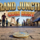 A viral, hyper-realistic YouTube thumbnail with a vibrant spring community event atmosphere. The background is a bustling downtown street scene with brick buildings, shop fronts, and clear blue Colorado skies with warm afternoon lighting. The composition uses a low angle shot to focus on the main subject: a gleaming golden commemorative coin resting on vintage brick pavement, with blurred silhouettes of families searching in the background. Image size should be 3:2. The image features massive 3D typography with strict hierarchy: The Primary Text reads exactly: 'GRAND JUNCTION'. This text is massive, the largest element in the frame, rendered in polished gold metallic material with engraved texture to look like a high-budget 3D render. The Secondary Text reads exactly: 'COIN HUNT'. This text is significantly smaller, positioned below the main text. It features a thick, distinct orange and red gradient border/outline (sticker style) with a playful adventure font to contrast against the background. Make sure text 2 is always different theme, style, effect and border compared to text 1. The text materials correspond to the story's concept. Crucial Instruction: There is absolutely NO other text, numbers, watermarks, or subtitles in this image other than these two specific lines. 8k, Unreal Engine 5, cinematic render.