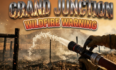 A viral, hyper-realistic YouTube thumbnail with a dramatic emergency response atmosphere. The background is a sun-scorched Colorado backyard with charred fence posts, swirling smoke, and a deep orange-amber gradient sky suggesting late morning heat. Harsh sunlight creates dramatic shadows while ember particles float in the air. The composition uses a low angle shot to emphasize urgency, focusing on the main subject: a professional fire hose nozzle spraying water in a powerful arc, with water droplets catching golden sunlight against the smoky background. Image size should be 3:2. The image features massive 3D typography with strict hierarchy: The Primary Text reads exactly: 'GRAND JUNCTION'. This text is massive, the largest element in the frame, rendered in scorched metallic chrome with flickering fire reflections and heat distortion effects to look like a high-budget 3D render. The Secondary Text reads exactly: 'WILDFIRE WARNING'. This text is significantly smaller, positioned below the main text. It features a thick, distinct bright red and yellow gradient border/outline in bold sticker style to contrast against the smoky background. Make sure text 2 is always different theme, style, effect and border compared to text 1. The text materials correspond to the story's concept of fire danger and emergency response. Crucial Instruction: There is absolutely NO other text, numbers, watermarks, or subtitles in this image other than these two specific lines. 8k, Unreal Engine 5, cinematic render.