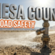 A viral, hyper-realistic YouTube thumbnail with a Utilitarian atmosphere. The background is a dusty rural highway shoulder with bright midday sunlight. The composition uses a Macro angle to focus on the main subject: a heavy duty industrial metal sprayer nozzle misting the ground. Image size should be 3:2. The image features massive 3D typography with strict hierarchy: The Primary Text reads exactly: 'MESA COUNTY'. This text is massive, the largest element in the frame, rendered in brushed steel material to look like a high-budget 3D render. The Secondary Text reads exactly: 'ROAD SAFETY'. This text is significantly smaller, positioned below the main text. It features a thick, bright orange color border outline sticker style to contrast against the background. Make sure text 2 is always different theme, style, effect and border compared to text 1.