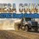 A viral, hyper-realistic YouTube thumbnail with a professional municipal infrastructure atmosphere. The background is a dusty Colorado dirt road stretching toward distant mountains with golden afternoon sunlight and swirling dust particles suspended in the air. The composition uses a low-angle camera perspective to focus on the main subject: a large metal road treatment tanker truck with magnesium chloride spraying equipment visible. The image features massive 3D typography with strict hierarchy: The Primary Text reads exactly: "MESA COUNTY". This text is massive, the largest element in the frame, rendered in shimmering chrome and metallic gold material to look like a high-budget 3D render with real-time reflections. The Secondary Text reads exactly: "275K Dust Solution". This text is significantly smaller, positioned below the primary text, featuring a thick bright blue neon border with a sticker-style outline that contrasts sharply against the dusty background. The text materials correspond to the professional infrastructure concept. Crucial Instruction: There is absolutely no other text, numbers, watermarks, or subtitles in this image other than these two specific lines. 8k, Unreal Engine 5, cinematic render.