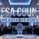 A viral, hyper-realistic YouTube thumbnail with a Sleek and Professional atmosphere. The background is a Modern Medical Gala Hall with Soft Blue and White Ambient Lighting. The composition uses a Low Angle to focus on the main subject: a Glass Crystal Health Award Trophy reflecting the lights. Image size should be 3:2. The image features massive 3D typography with strict hierarchy: The Primary Text reads exactly: 'MESA COUNTY'. This text is massive, the largest element in the frame, rendered in Chrome to look like a high-budget 3D render. The Secondary Text reads exactly: 'HEALTH HEROES'. This text is significantly smaller, positioned below the main text. It features a thick, distinct Blue sticker style border to contrast against the background. The text materials correspond to the story's concept. Crucial Instruction: There is absolutely NO other text, numbers, watermarks, or subtitles in this image other than these two specific lines. 8k, Unreal Engine 5, cinematic render.
