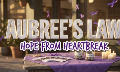 A viral, hyper-realistic image with a somber yet hopeful atmosphere blending community activism and memorial themes. The background is a softly lit bookstore exterior with warm golden hour lighting casting long shadows, featuring purple ribbons (domestic violence awareness symbol) gently floating in the breeze. The composition uses an eye-level medium shot to focus on the main subject: a weathered wooden fundraising table draped with a purple awareness cloth, topped with flickering memorial candles, donation jars filled with bills and coins, and awareness brochures fanned across the surface. Image size should be 3:2. The image features massive 3D typography with strict hierarchy: The Primary Text reads exactly: 'AUBREE'S LAW'. This text is massive, the largest element in the frame, rendered in polished marble with gold veining and soft ethereal glow to look like a high-budget 3D render. The Secondary Text reads exactly: 'HOPE FROM HEARTBREAK'. This text is significantly smaller, positioned below the main text. It features a thick, distinct purple border with white outline (sticker style) in a handwritten font that contrasts against the background. Make sure text 2 is always different theme, style, effect and border compared to text 1. The text materials correspond to the story's concept of memorial and legislative victory. Crucial Instruction: There is absolutely NO other text, numbers, watermarks, or subtitles in this image other than these two specific lines. 8k, Unreal Engine 5, cinematic render.