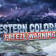 A viral, hyper-realistic YouTube thumbnail with a dramatic mountain weather atmosphere. The background is a sweeping Colorado mountain landscape at dusk with dark storm clouds rolling over snow-capped peaks, illuminated by dramatic purple and blue lighting with occasional lightning strikes in the distance. The composition uses a wide-angle low angle to focus on the main subject: massive snowflakes falling over a detailed topographic mountain range with a thermometer showing freezing temperatures embedded in fresh snow. Image size should be 3:2. The image features massive 3D typography with strict hierarchy: The Primary Text reads exactly: 'WESTERN COLORADO'. This text is massive, the largest element in the frame, rendered in Icy Chrome with realistic snow accumulation on the letters to look like a high-budget 3D render. The Secondary Text reads exactly: 'FREEZE WARNING'. This text is significantly smaller, positioned below the main text. It features a thick, distinct red and white border/outline (sticker style) with a frost effect to contrast against the background. Make sure text 2 is always different theme, style, effect and border compared to text 1. The text materials correspond to the story's concept. Crucial Instruction: There is absolutely NO other text, numbers, watermarks, or subtitles in this image other than these two specific lines. 8k, Unreal Engine 5, cinematic render.